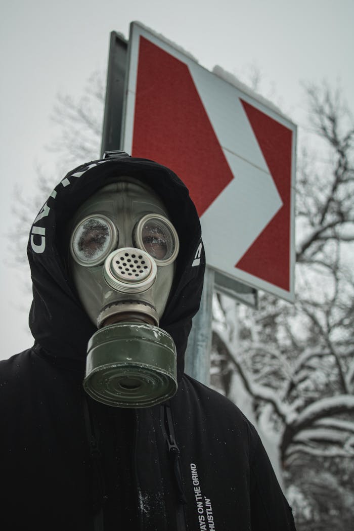 Individual wearing a gas mask stands next to a road sign amidst snow-covered trees.